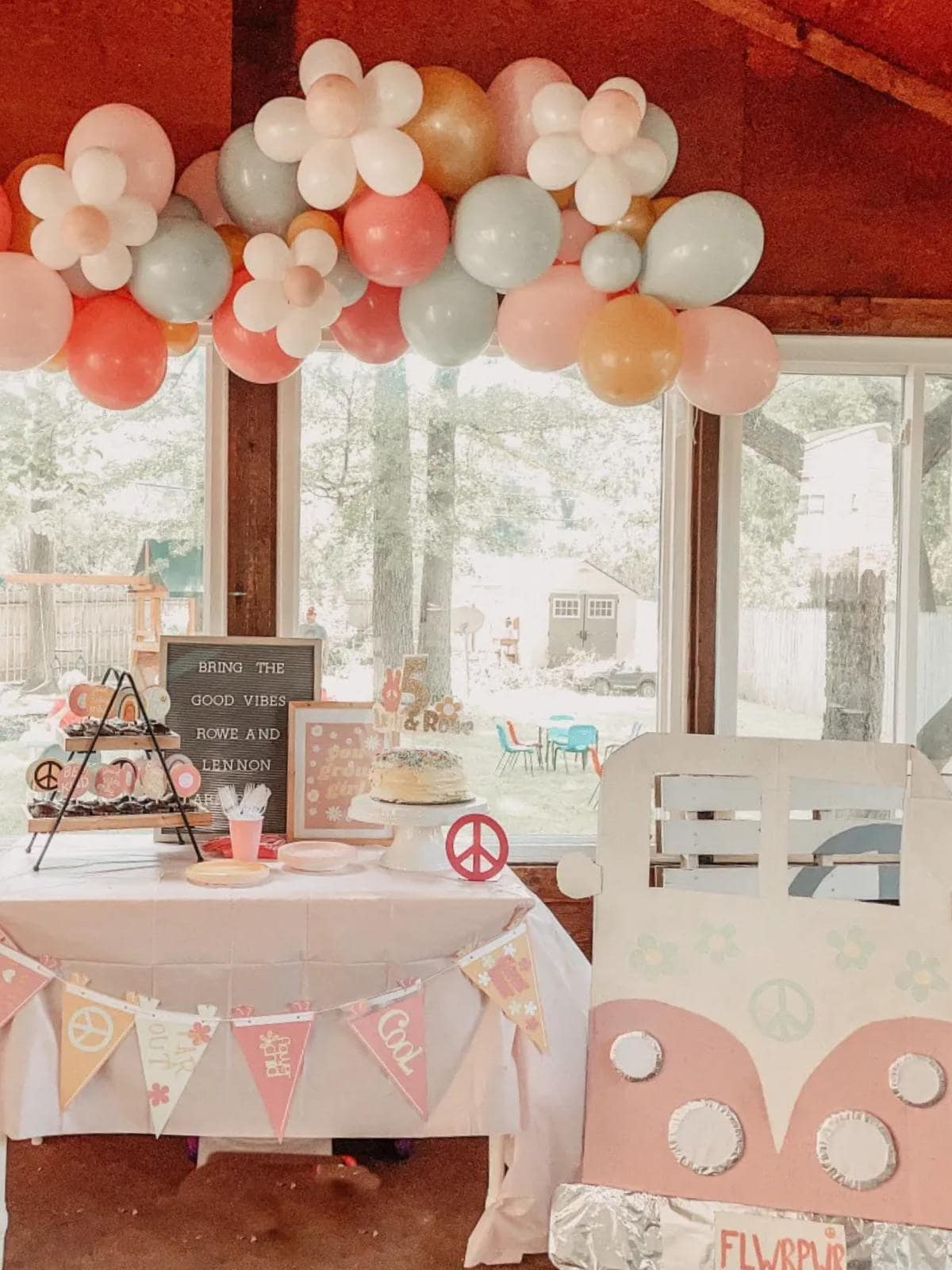 A pastel-themed birthday party setup features a table with a cake, peace sign décor, a letter board, and a pink and white van cutout. Above are colorful balloons shaped like flowers and orbs. The space has large windows and outdoor views.