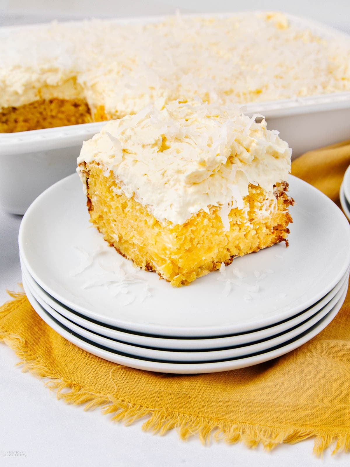 A slice of yellow cake with creamy frosting and shredded coconut on top sits on a stack of white plates, with a larger dish of the same cake in the background on a yellow cloth.