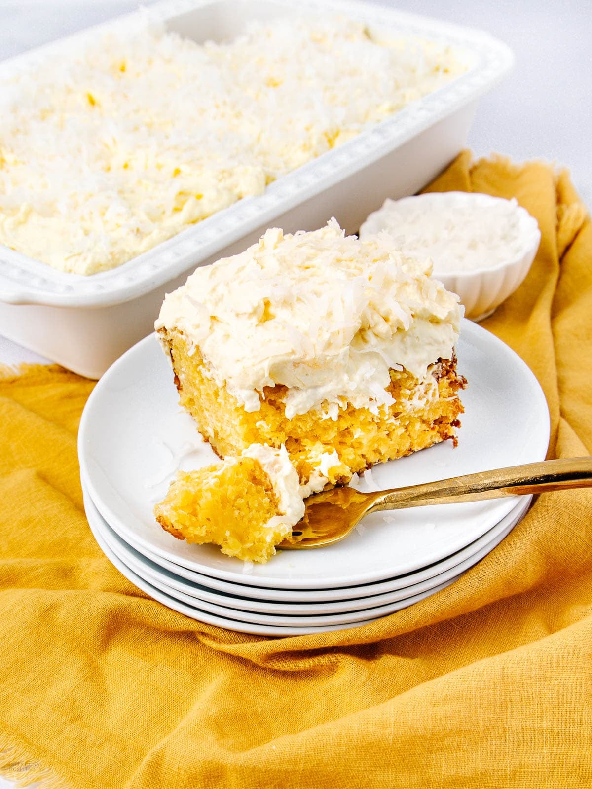 A square slice of coconut cake with creamy frosting sits on a white plate, topped with shredded coconut. A fork holds a bite of cake. More cake and coconut are in dishes in the background, all on a yellow cloth.