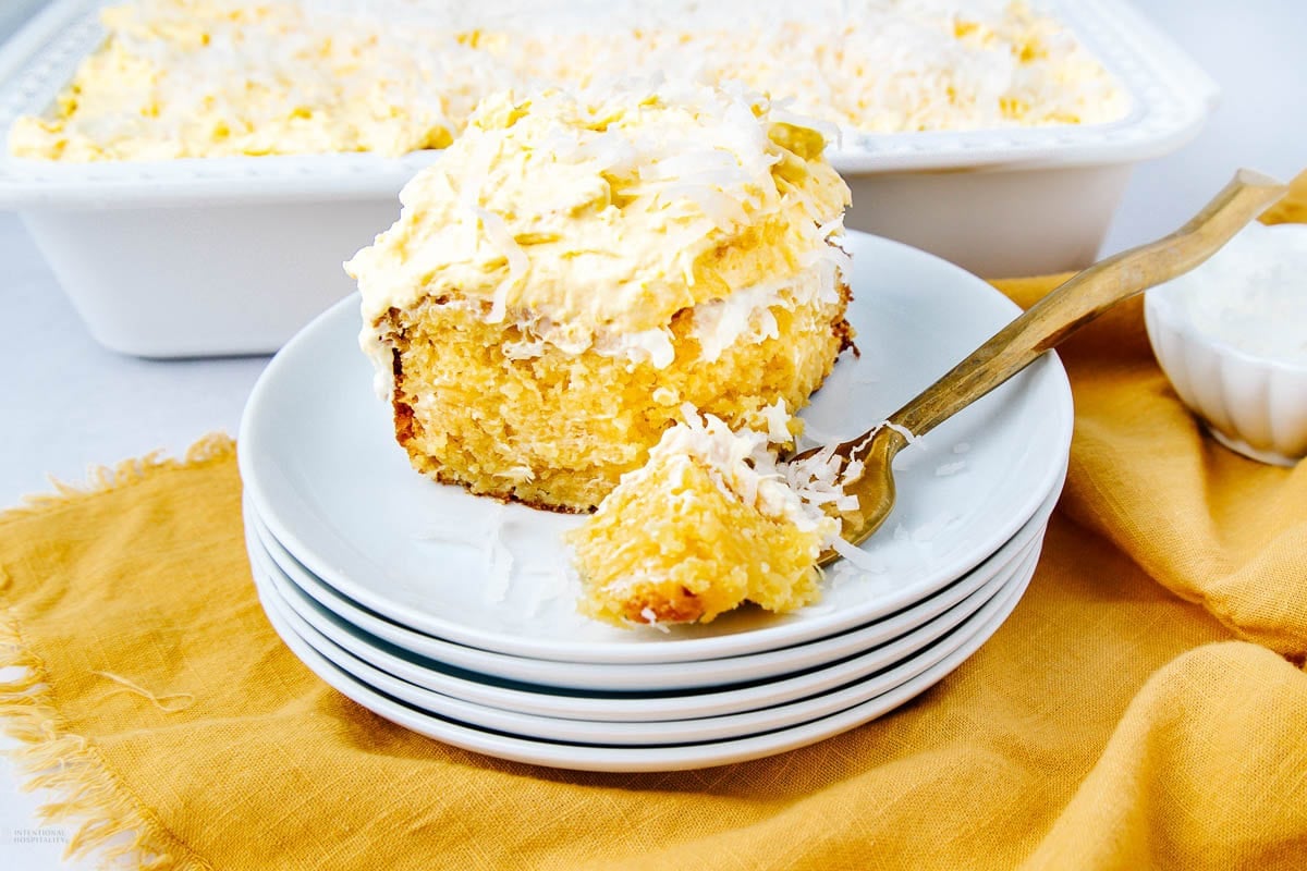 A slice of coconut cake with fluffy frosting and shredded coconut on top sits on a white plate with a fork, in front of a dish of more cake, on a yellow cloth.