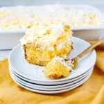 A slice of moist yellow cake topped with creamy frosting and shredded coconut sits on a stack of white plates, with a fork beside it. A large dish of the same cake is in the background on a yellow cloth.