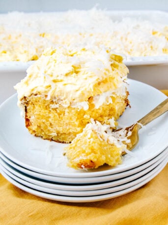 A slice of moist yellow cake topped with creamy frosting and shredded coconut sits on a stack of white plates, with a fork beside it. A large dish of the same cake is in the background on a yellow cloth.