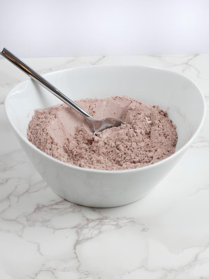 A white bowl filled with light brown baking mix or powder sits on a marble surface. A metal spoon is partially buried in the powder, resting along the side of the bowl.
