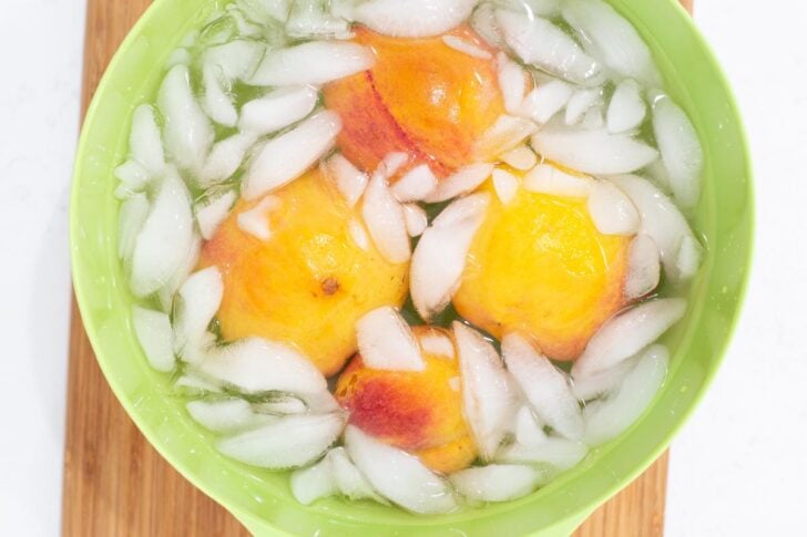 Three peaches are submerged in a green bowl filled with ice water and ice cubes, sitting on a wooden surface.