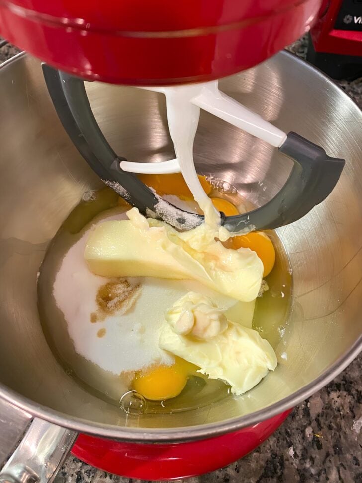A close-up of a red stand mixer bowl with butter, eggs, sugar, and vanilla before mixing, ready to be blended for baking on a granite countertop.