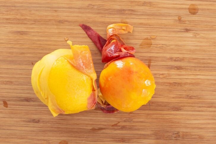 Two peaches partly peeled with their skins curling off, placed on a wooden cutting board.