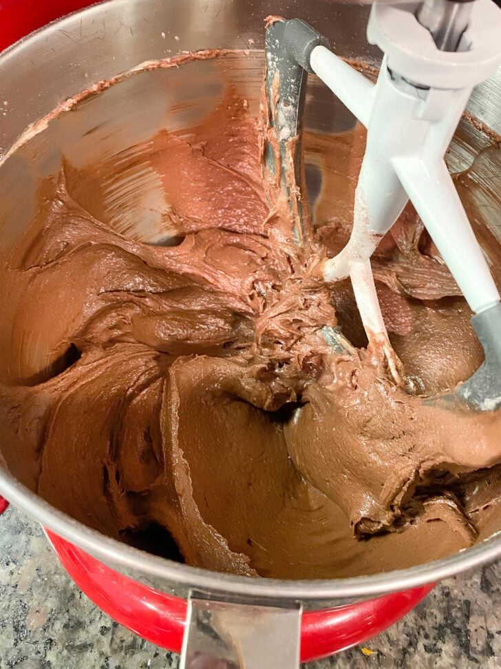 A close-up of chocolate cake batter being mixed in a stand mixer with a paddle attachment. The batter is thick and creamy, and the mixing bowl is silver with a red base.