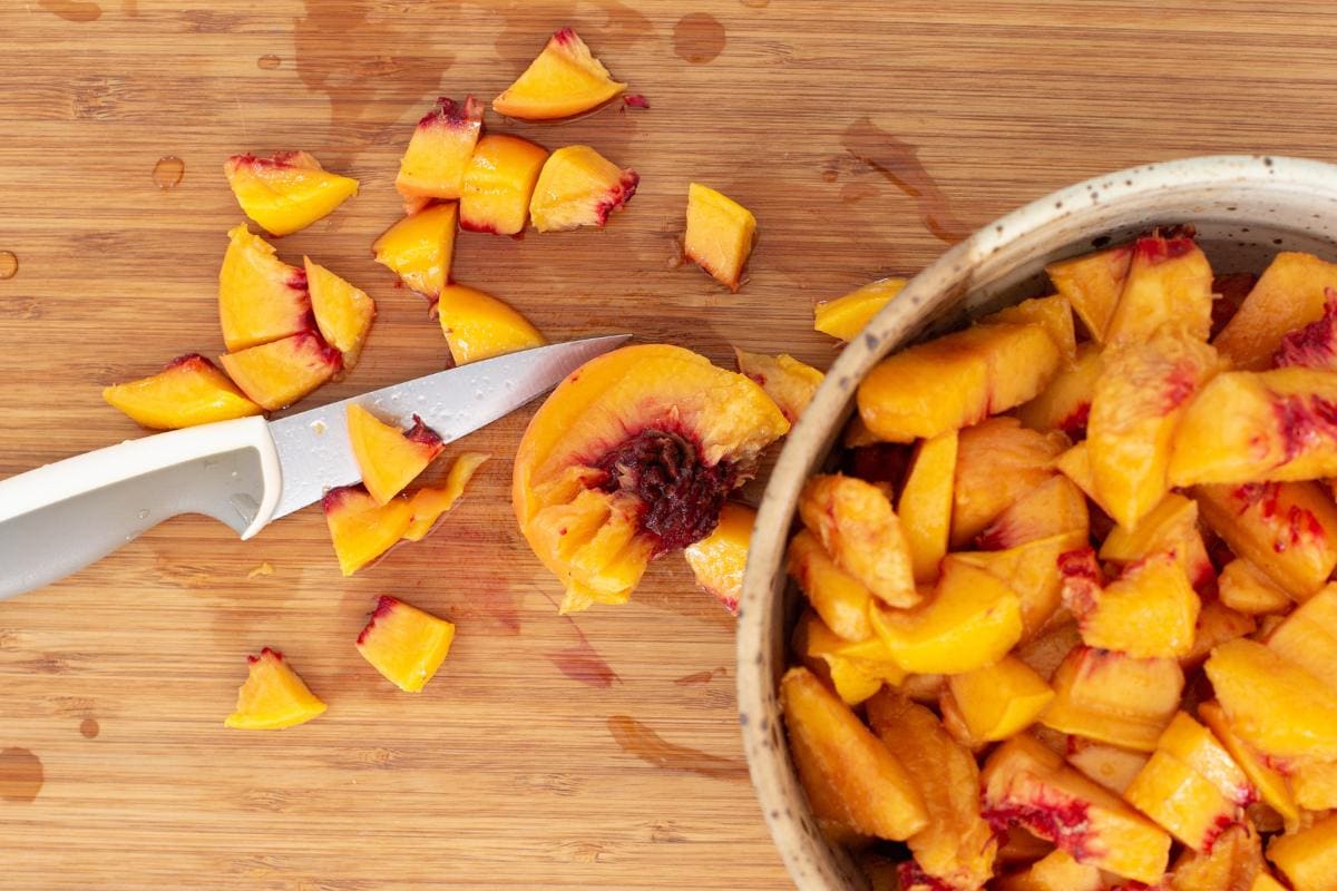 A knife and chopped peaches are on a wooden cutting board, with a bowl filled with more chopped peaches to the right.