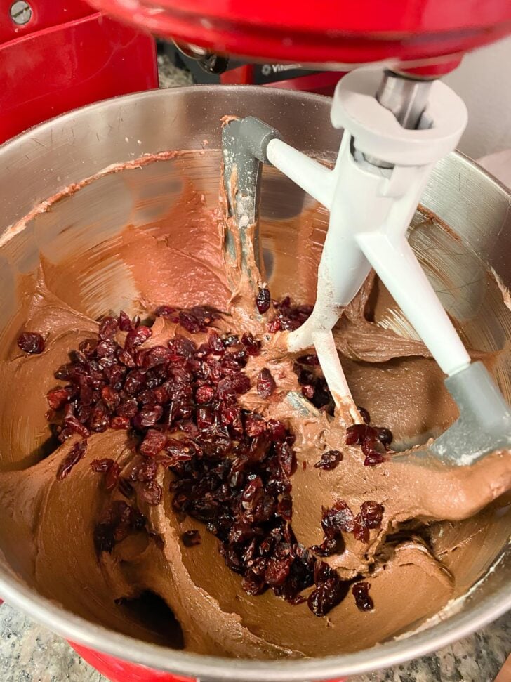 A close-up of a stand mixer bowl filled with chocolate batter, dried cranberries, and a paddle attachment, ready for mixing.