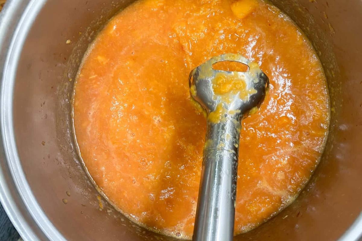 An immersion blender is in a pot of orange puree, likely a soup or sauce, with a smooth and slightly chunky texture.