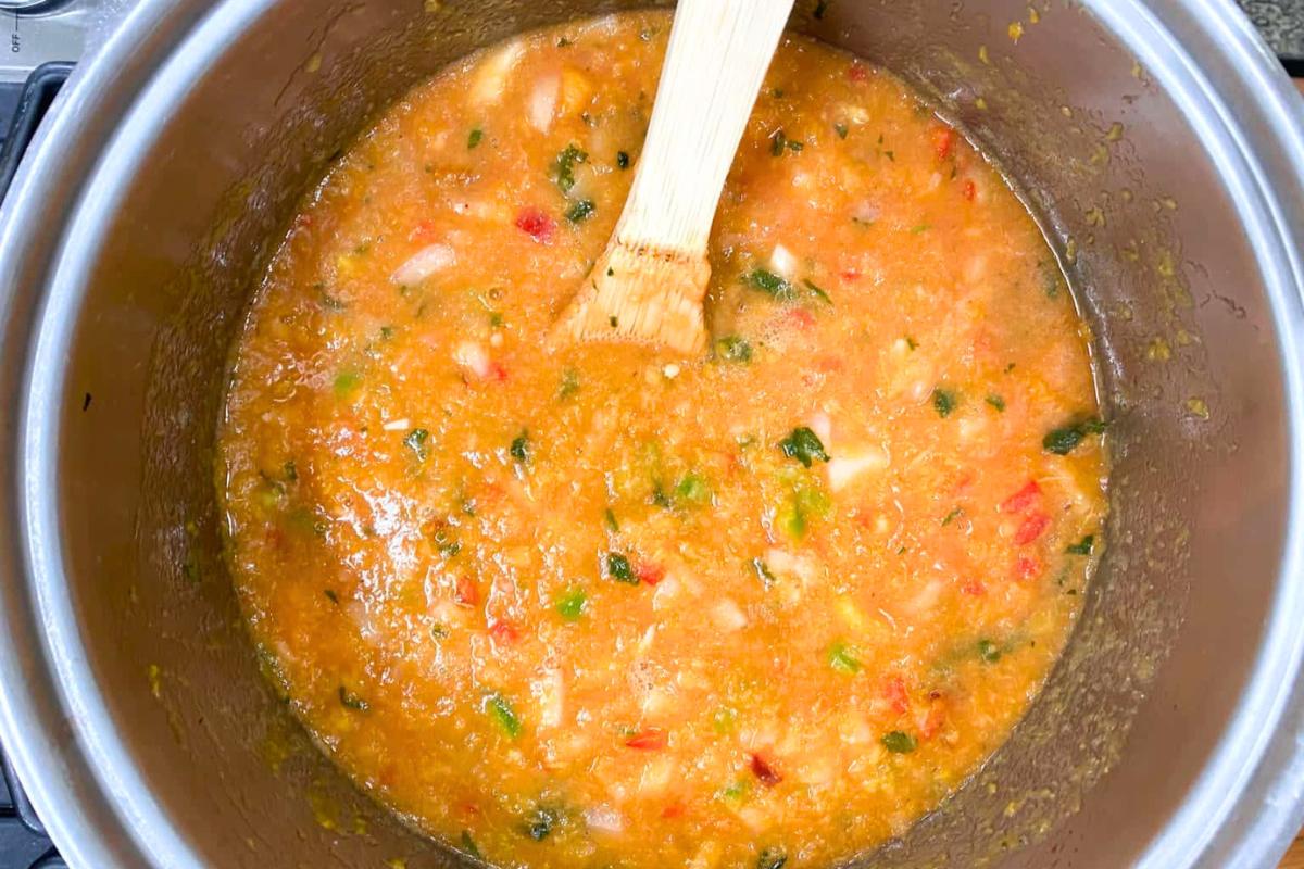 A pot of blended tomato sauce with visible chunks of onions and herbs is being stirred with a wooden spatula. The sauce has a vibrant orange-red color and a slightly chunky texture.