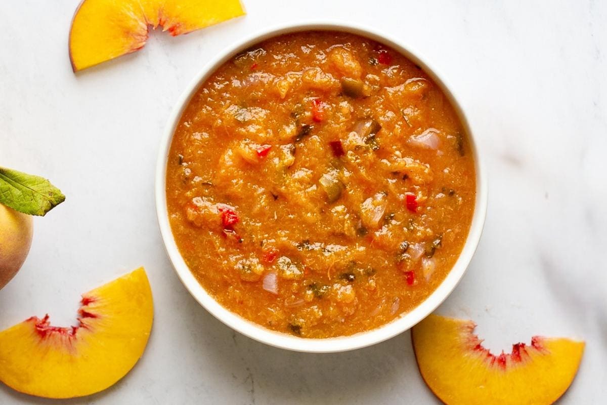 A bowl of chunky peach salsa sits on a white surface, surrounded by three fresh peach slices and part of a whole peach with a leaf.
