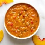 A white bowl filled with chunky peach salsa, featuring visible pieces of red pepper and onion, sits on a white marble surface with peach slices nearby.