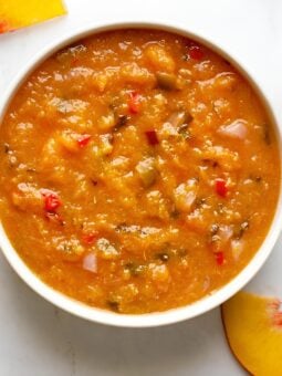A white bowl filled with chunky peach salsa, featuring visible pieces of red pepper and onion, sits on a white marble surface with peach slices nearby.