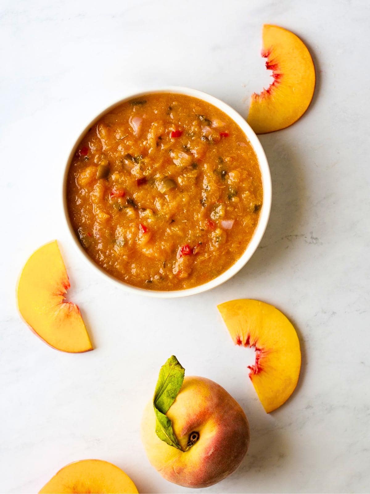 A bowl of chunky peach salsa sits on a white surface, surrounded by peach slices and a whole peach with a green leaf.