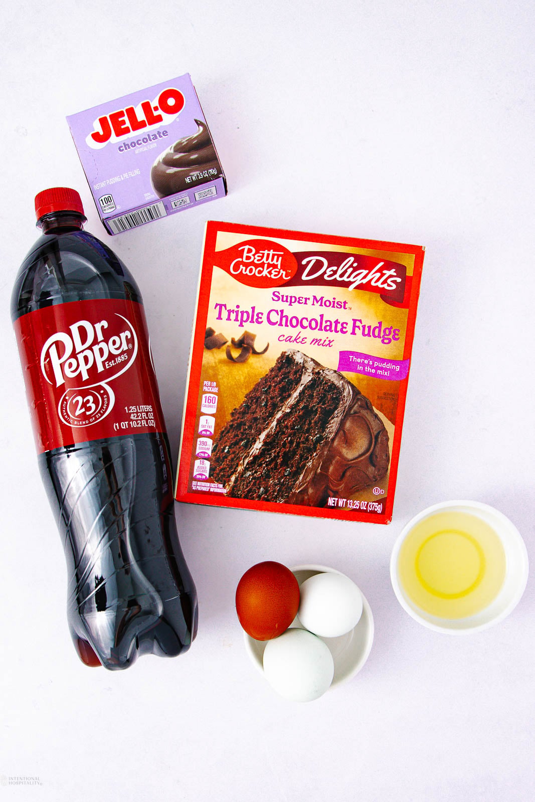 A bottle of Dr Pepper, a box of chocolate Jello pudding, a box of Betty Crocker triple chocolate fudge cake mix, three eggs, and a bowl of oil are arranged on a white surface.