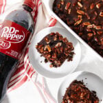 A bottle of Dr Pepper next to two plates with chocolate pecan cake slices, and a baking dish with more cake, all on a white surface with a red-striped towel.