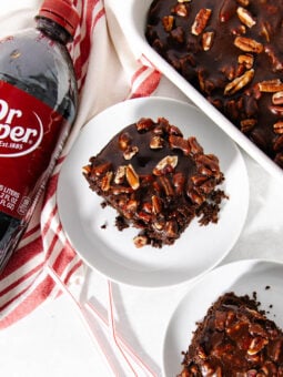 A bottle of Dr Pepper next to two plates with chocolate pecan cake slices, and a baking dish with more cake, all on a white surface with a red-striped towel.