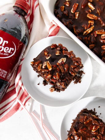 A bottle of Dr Pepper next to two plates with chocolate pecan cake slices, and a baking dish with more cake, all on a white surface with a red-striped towel.