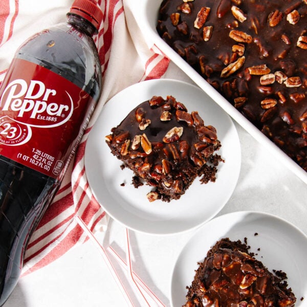 A bottle of Dr Pepper next to two plates with chocolate pecan cake slices, and a baking dish with more cake, all on a white surface with a red-striped towel.