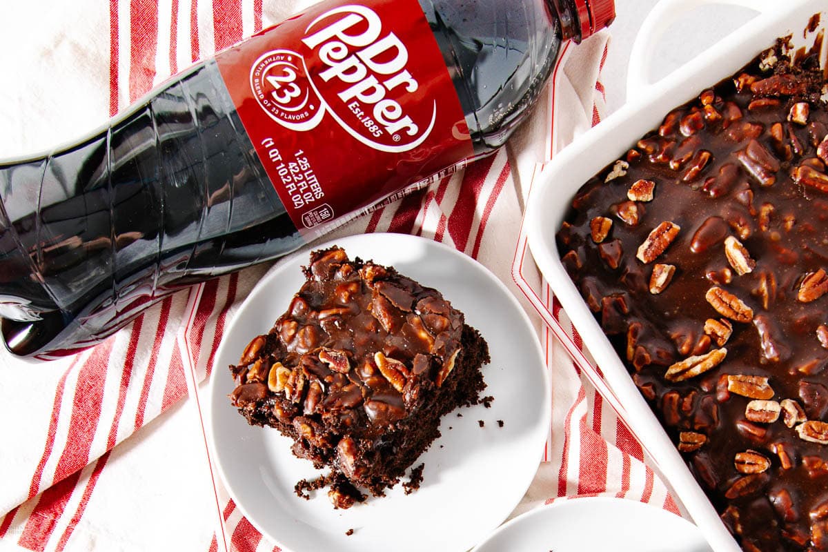 A bottle of Dr Pepper lies next to a plate with a chocolate pecan brownie and a baking dish with more brownies on a red and white striped towel.