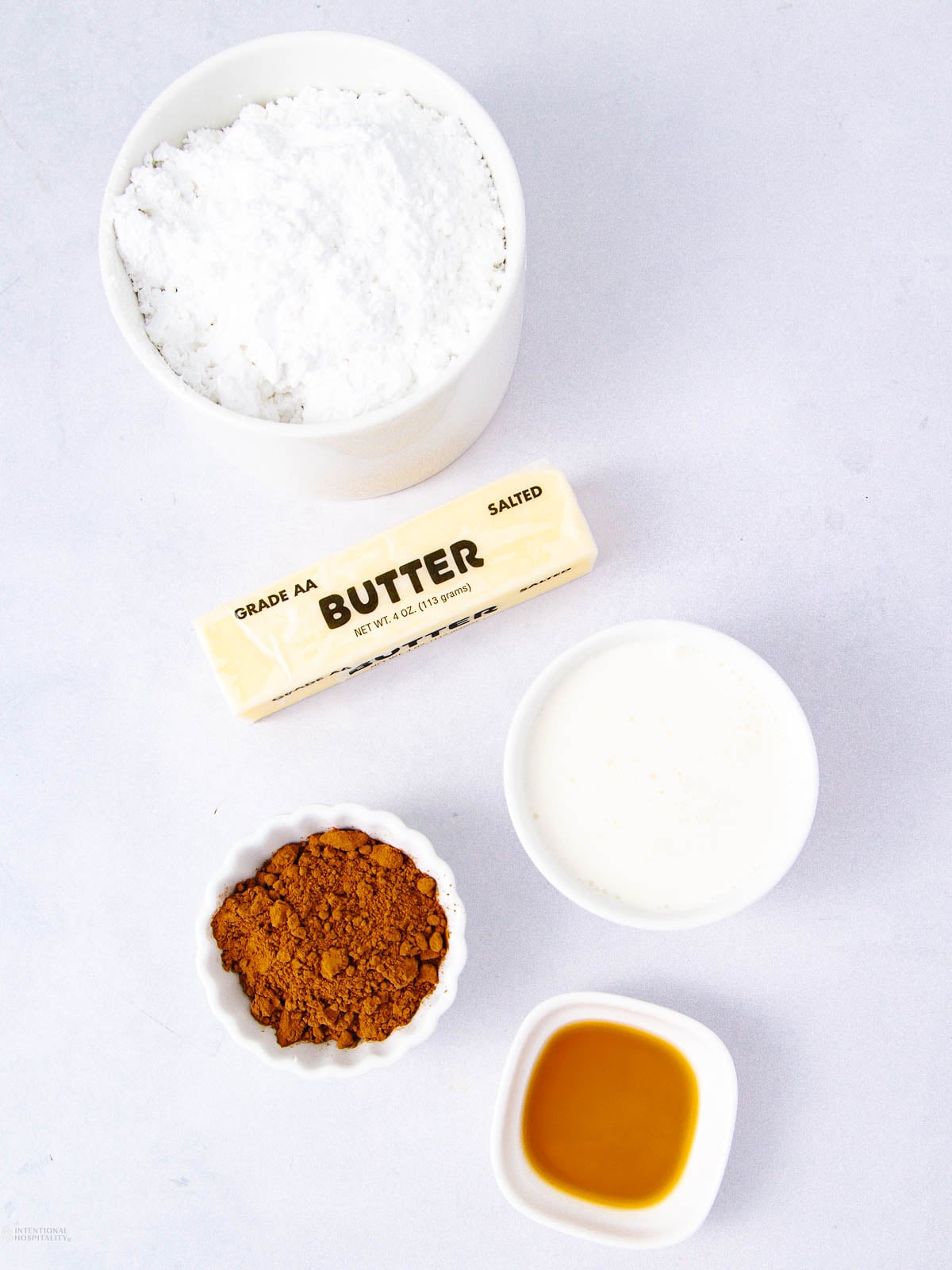 Five baking ingredients on a white surface: a bowl of powdered sugar, a stick of butter, a small bowl of milk, a dish of cocoa powder, and a dish of vanilla extract.