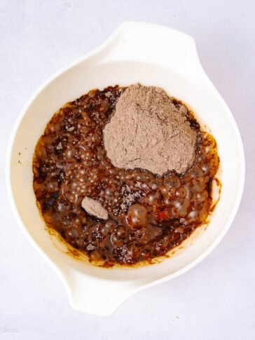 A white mixing bowl with a blend of brown liquid, small translucent tapioca pearls, and a mound of dry chocolate powder on top, set against a light background.