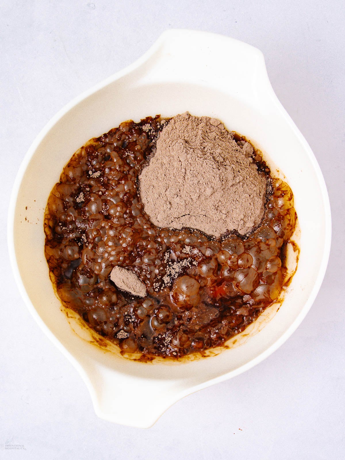 A white mixing bowl with a blend of brown liquid, small translucent tapioca pearls, and a mound of dry chocolate powder on top, set against a light background.