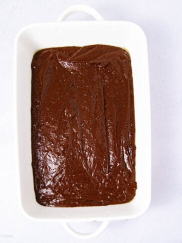 A white baking dish filled with smooth, unbaked chocolate brownie batter, viewed from above on a white background.