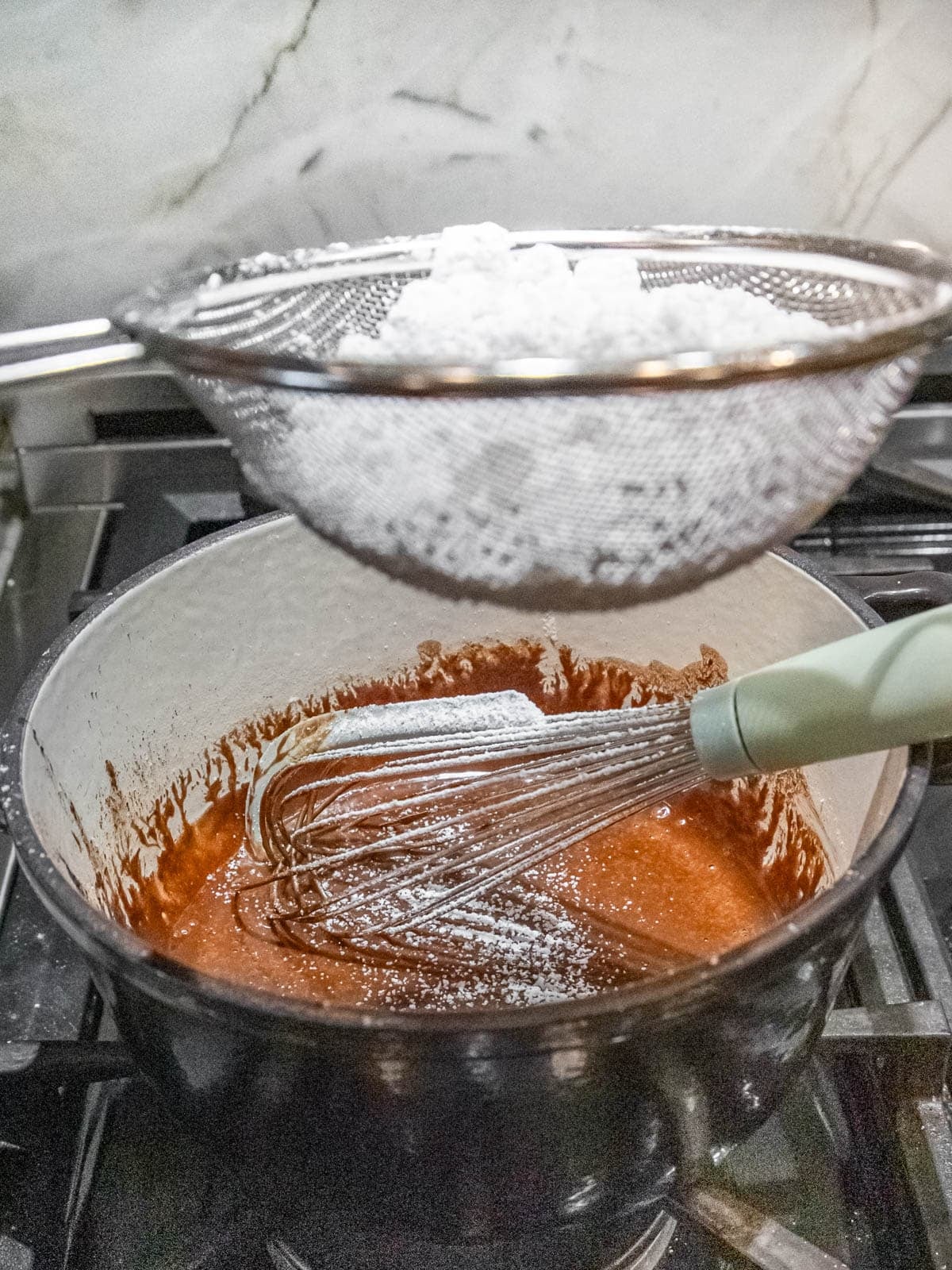 Powdered sugar is sifted into a pot of chocolate batter on a stovetop, with a whisk resting inside the pot and a mesh strainer held above.