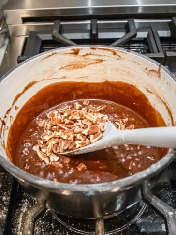 Chopped pecans are being stirred into a pot of bubbling chocolate mixture on a stove, using a white spoon.