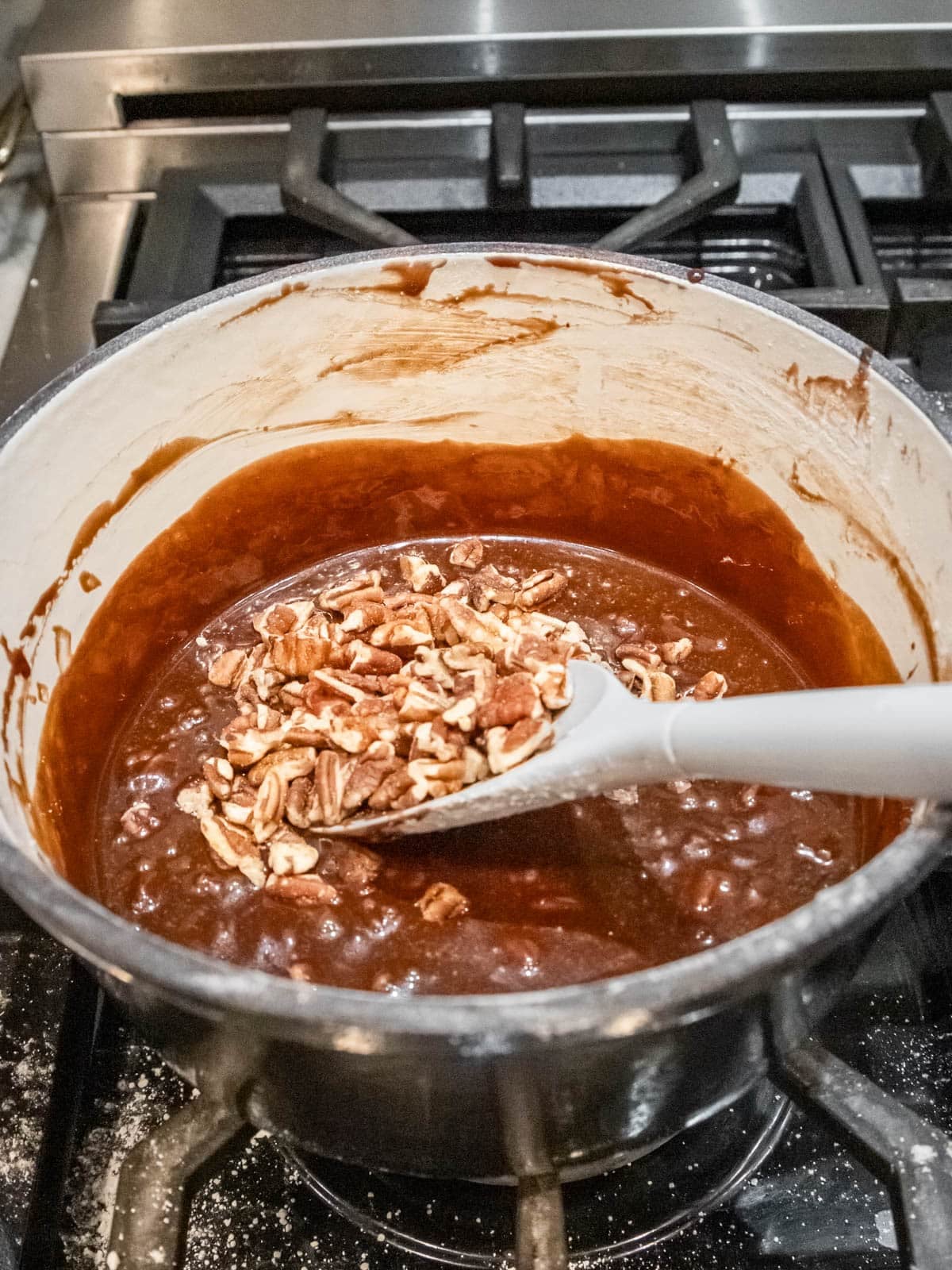 Chopped pecans are being stirred into a pot of bubbling chocolate mixture on a stove, using a white spoon.
