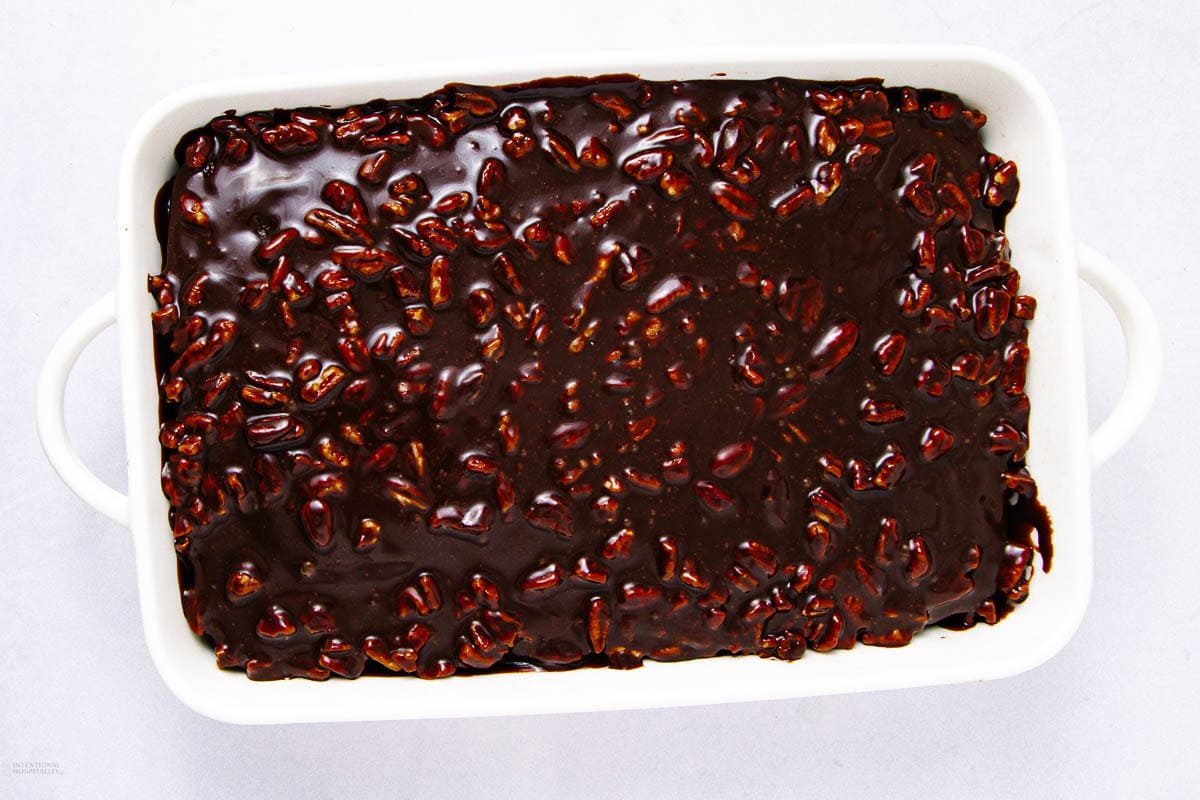 A rectangular white baking dish filled with chocolatey dessert topped with a glossy layer of chocolate and pecans.