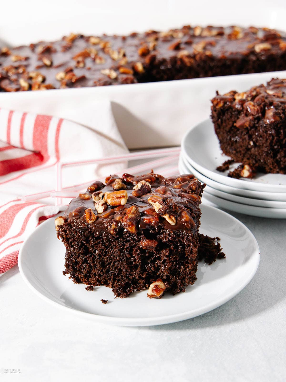 A slice of chocolate sheet cake with pecans and chocolate glaze sits on a white plate. More cake and plates are in the background, with a red and white striped towel nearby.