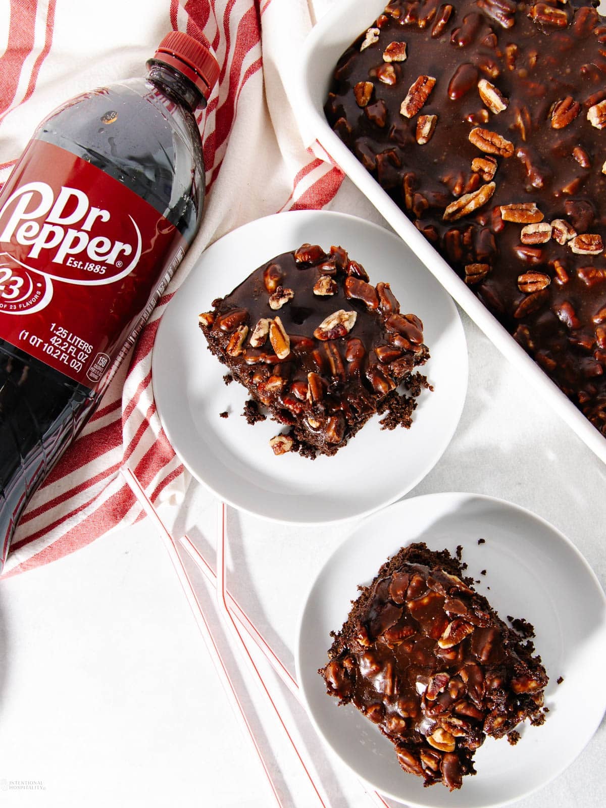 Two plates with chocolate pecan cake slices next to a pan of cake and a bottle of Dr Pepper, on a white surface with a red-striped kitchen towel.