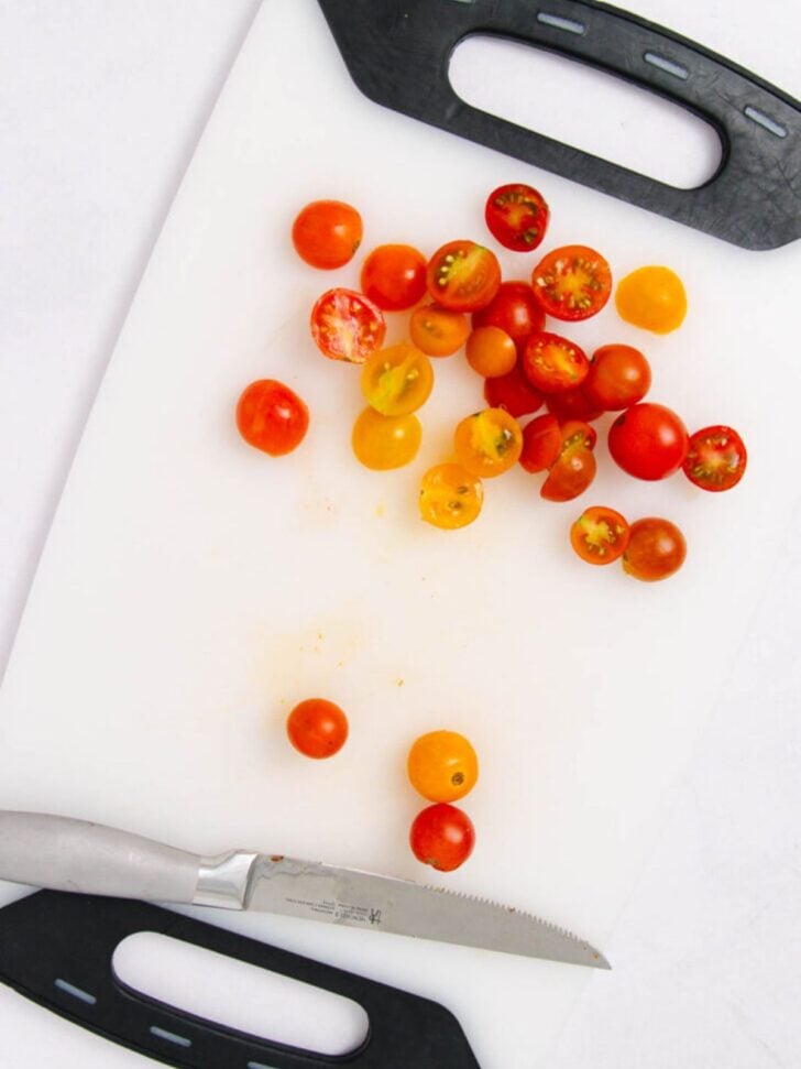 A white cutting board with a mix of whole and halved red and yellow cherry tomatoes, perfect for preparing a marinated veggie salad, and a serrated knife with a silver handle placed at the bottom.