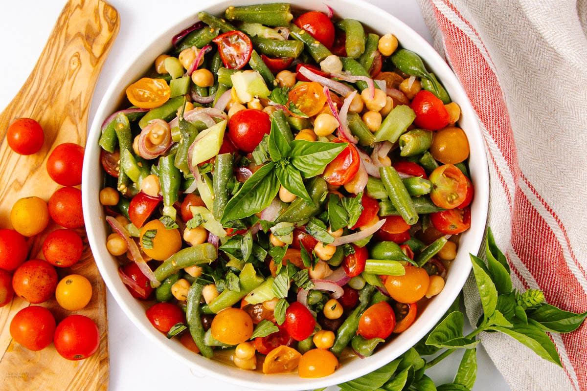 A bowl of colorful salad with cherry tomatoes, green beans, chickpeas, cucumbers, red onions, and fresh basil, placed next to loose tomatoes and a striped towel.