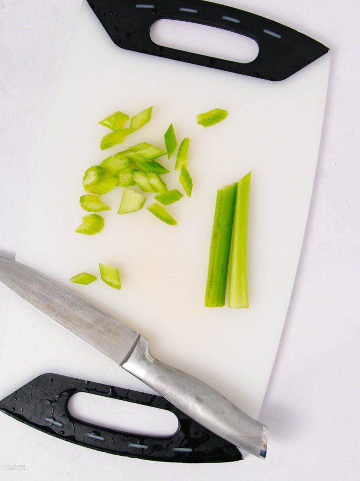 A cutting board with a chef’s knife, sliced and whole celery stalks, and celery pieces scattered on the surface.