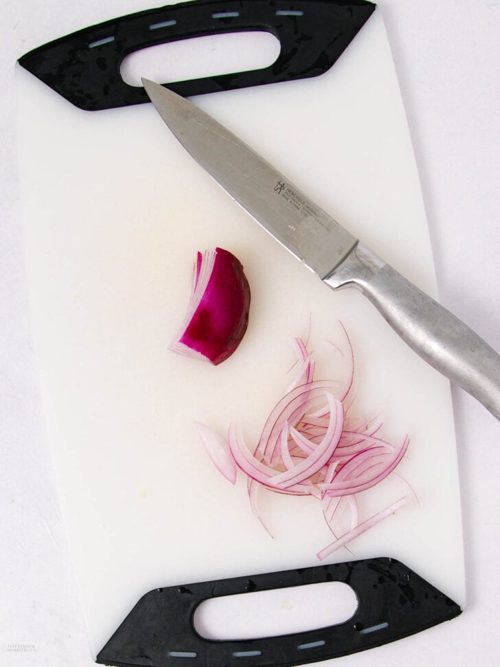 A stainless steel knife, sliced red onion, and a chunk of whole red onion rest on a white cutting board with black handles.