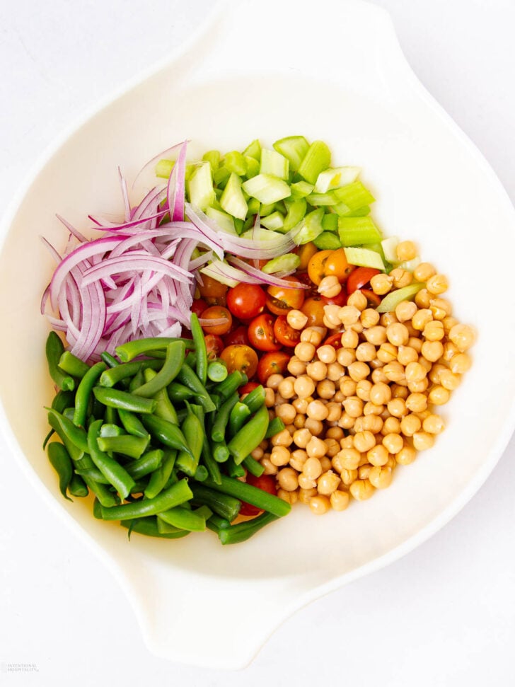 A white bowl with sliced red onion, diced celery, cherry tomatoes, chickpeas, and chopped green beans arranged in separate sections.