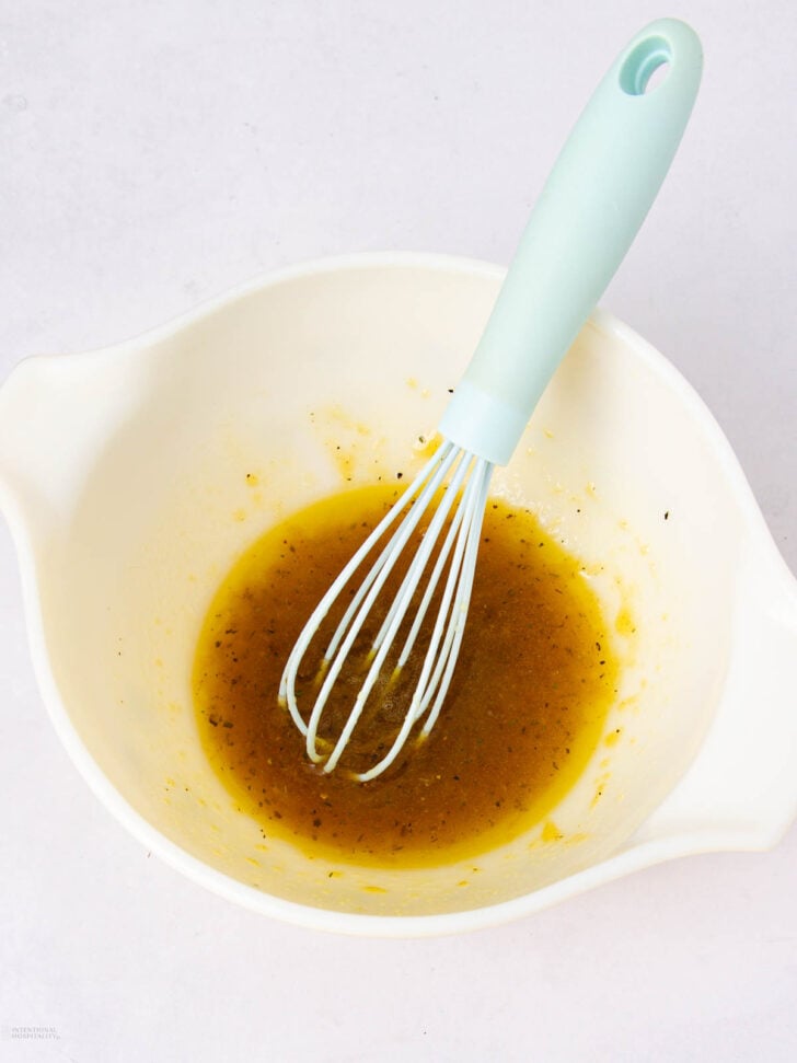 A white mixing bowl with yellow vinaigrette dressing and a light blue whisk inside, set on a white surface.