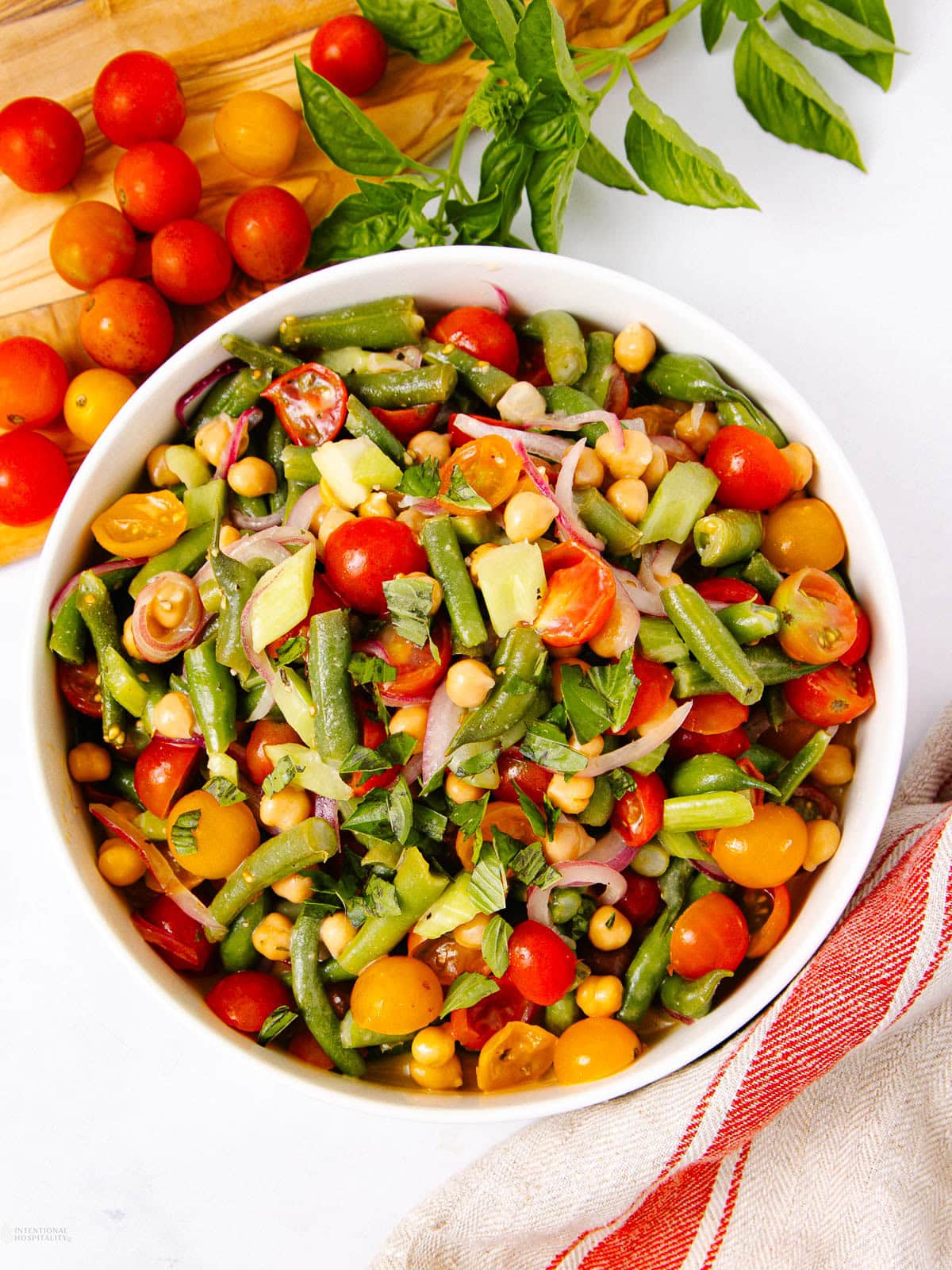 A bowl of colorful salad with green beans, cherry tomatoes, chickpeas, red onions, and fresh herbs on a white surface, with tomatoes and basil nearby.