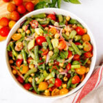 A bowl of colorful salad with green beans, chickpeas, cherry tomatoes, red onions, cucumber, and fresh herbs on a white surface, next to a red-striped towel.