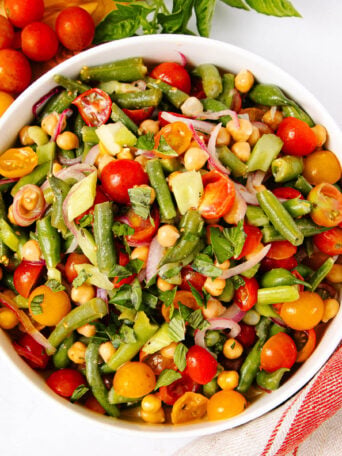A bowl of colorful salad with green beans, chickpeas, cherry tomatoes, red onions, cucumber, and fresh herbs on a white surface, next to a red-striped towel.
