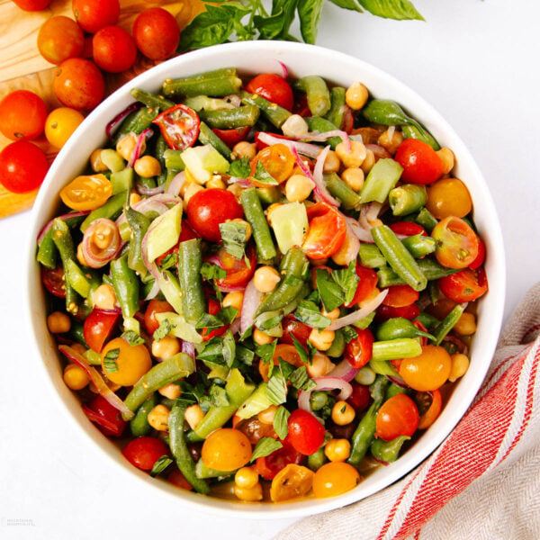 A bowl of colorful salad with green beans, chickpeas, cherry tomatoes, red onions, cucumber, and fresh herbs on a white surface, next to a red-striped towel.