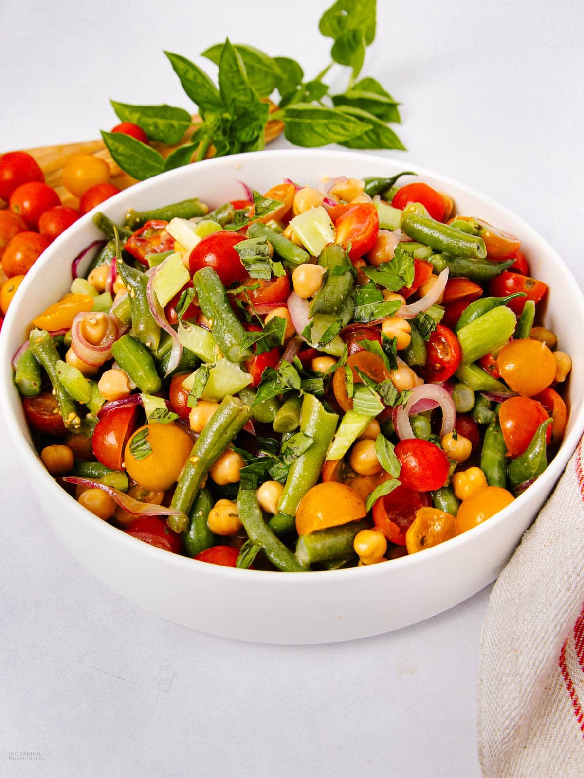 A white bowl filled with a colorful salad of green beans, cherry tomatoes, chickpeas, red onions, and herbs sits on a white surface. Fresh herbs and more tomatoes are in the background.
