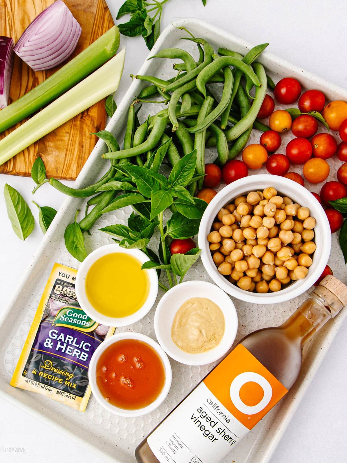 Tray with fresh green beans, cherry tomatoes, chickpeas, basil, celery, red onion, olive oil, hot sauce, Dijon mustard, balsamic vinegar, and a garlic & herb dressing packet arranged on a white surface.