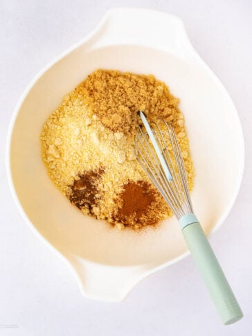 A white mixing bowl with cornmeal, brown sugar, cinnamon, and spices. A metal whisk with a light green handle rests inside the bowl on top of the dry ingredients.