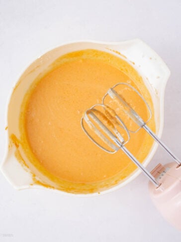 A white mixing bowl with orange batter and a pink electric hand mixer with two beaters resting inside. The bowl and mixer are on a white surface.