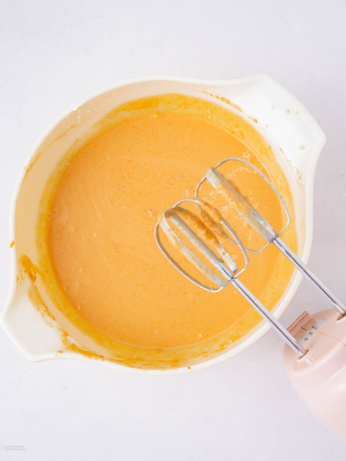 A white mixing bowl with orange batter and a pink electric hand mixer with two beaters resting inside. The bowl and mixer are on a white surface.
